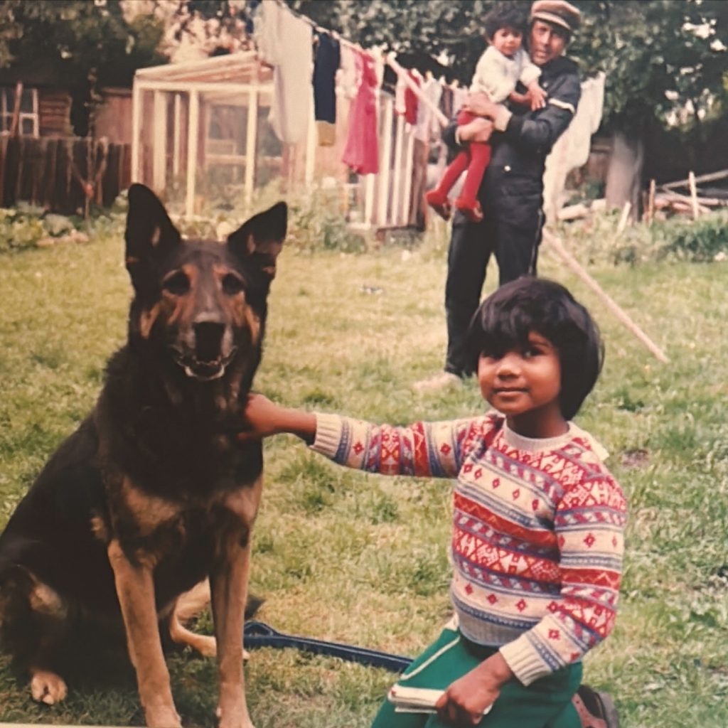 A young child (Nimi) with dark hair and a bright, patterned jumper kneels in a grassy garden next to a large German Shepherd dog, gently resting a hand on its back. In the background, a man in a dark uniform holds a baby while standing near a clothesline and wooden sheds. The scene has a nostalgic feel, hinting at a close family moment in a suburban garden.