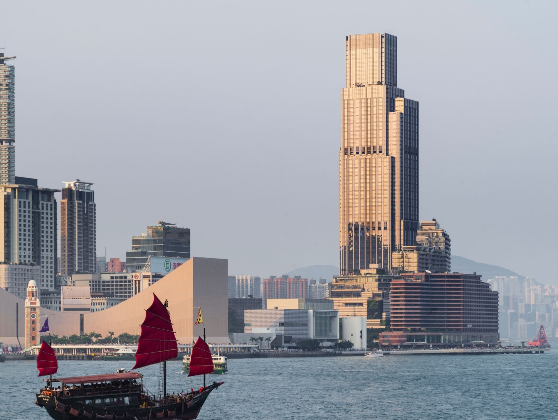 Waterfront view of Victoria Dockside in Hong Kong, with KPF’s Rosewood Tower catching the evening light. A traditional junk boat with red sails sails across the foreground.