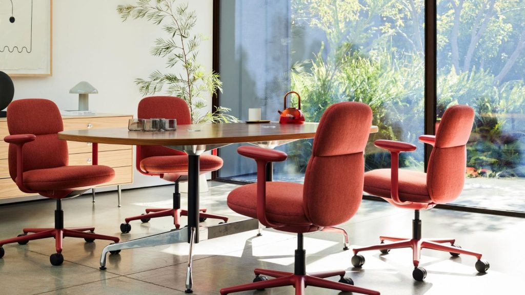 A modern meeting area with four red upholstered Asari chairs by Herman Miller arranged around a rectangular wooden table. The chairs feature curved backs, padded armrests, and five-point swivel bases with castors. The space is bright and airy, with large floor-to-ceiling glass windows revealing lush greenery outside. A red teapot and minimal decor sit atop the table. The background includes a low wooden sideboard, a lamp, and framed artwork on the wall, contributing to a warm, contemporary aesthetic.