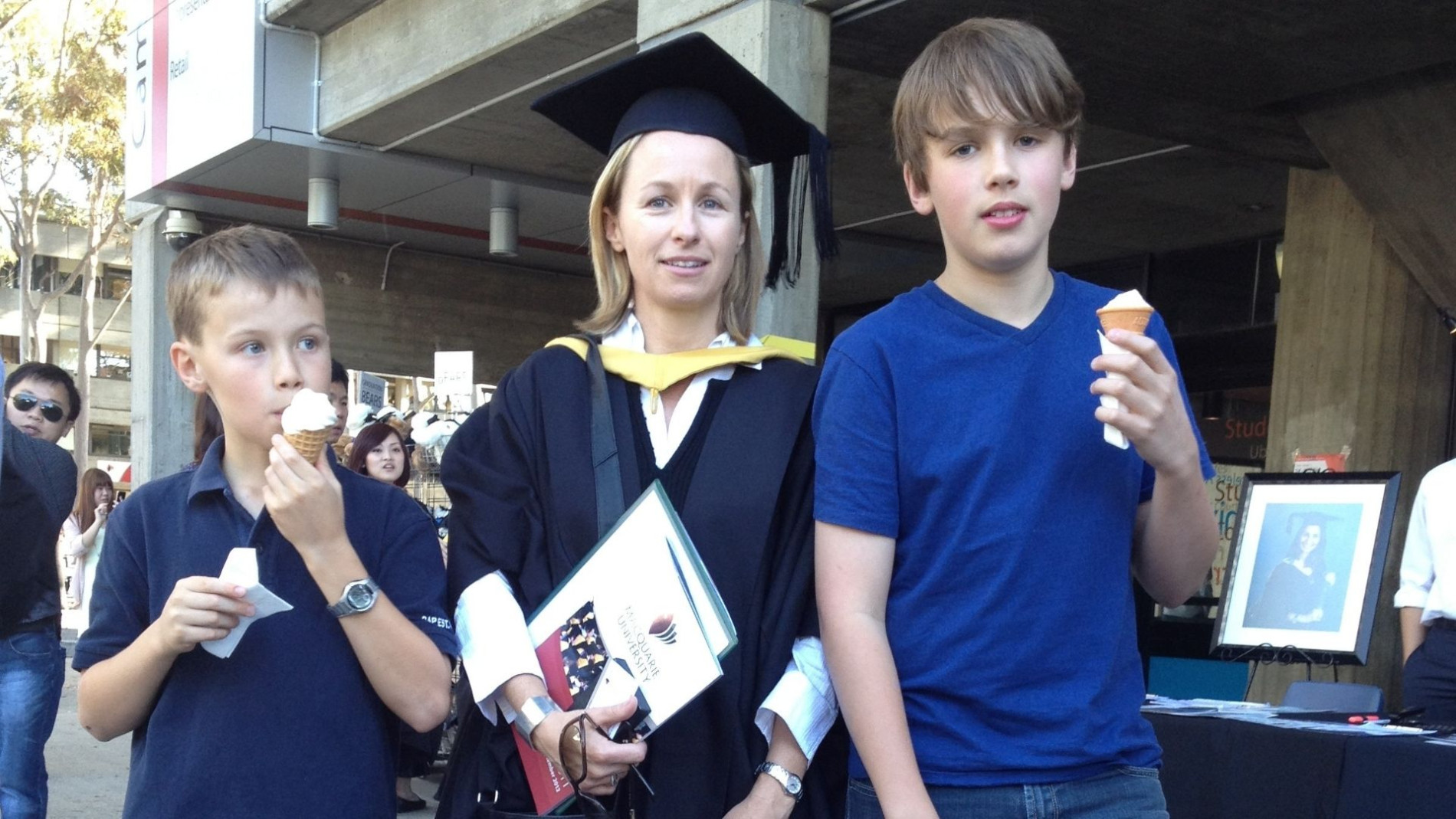 Kirsten in full grad regalia, holding her certificate. Her two boys stand next to her eating ice creams.