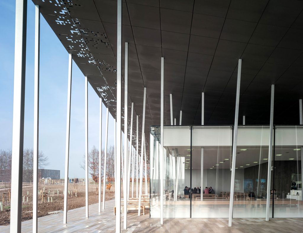 View from beneath the canopy of the Stonehenge Visitor Centre, designed by Denton Corker Marshall in 2013, showing slender angled columns, perforated roof panels, and floor-to-ceiling glass walls overlooking the surrounding landscape.