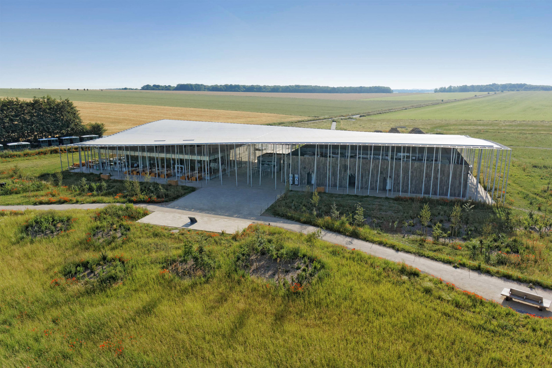 Aerial view of the Stonehenge Visitor Centre, designed by Denton Corker Marshall in 2013, featuring a flat roof supported by slender vertical columns, glazed walls, and surrounding grassland in the Wiltshire countryside.