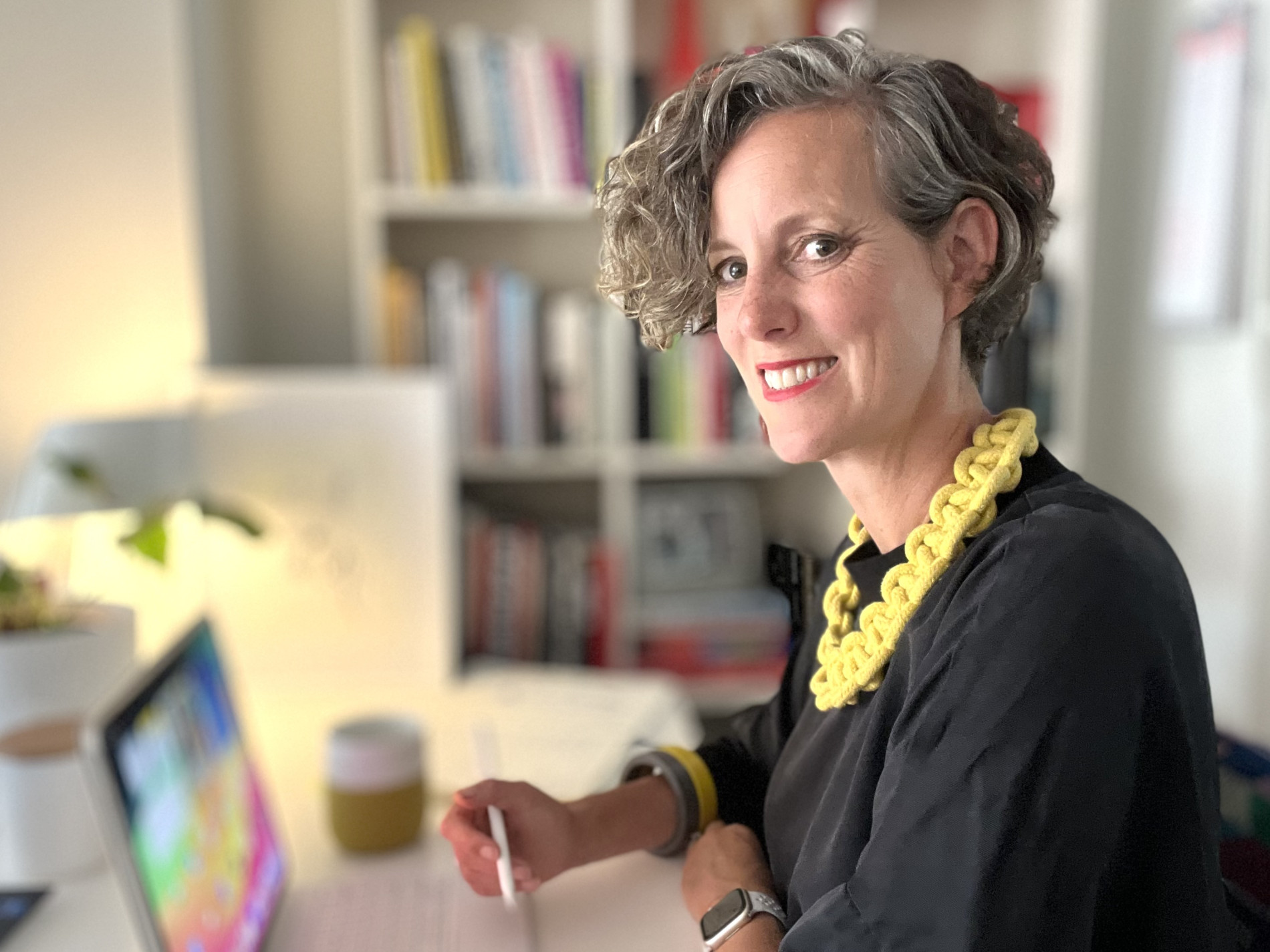 Angela Dapper seated at a desk with a digital tablet, smiling at the camera. She wears a black top with a chunky yellow necklace, with bookshelves blurred in the background.