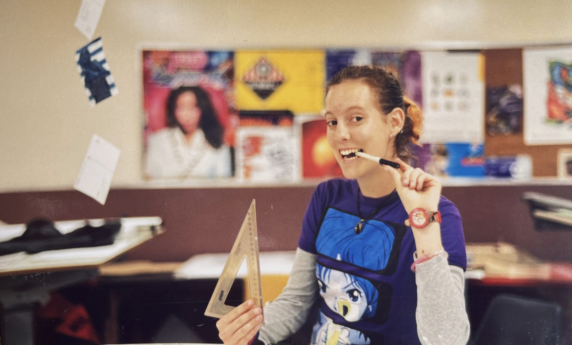 Angela Dapper at the University of Adelaide in 1997, smiling while holding a triangular ruler and pencil in a design studio, with posters and drafting tables in the background.