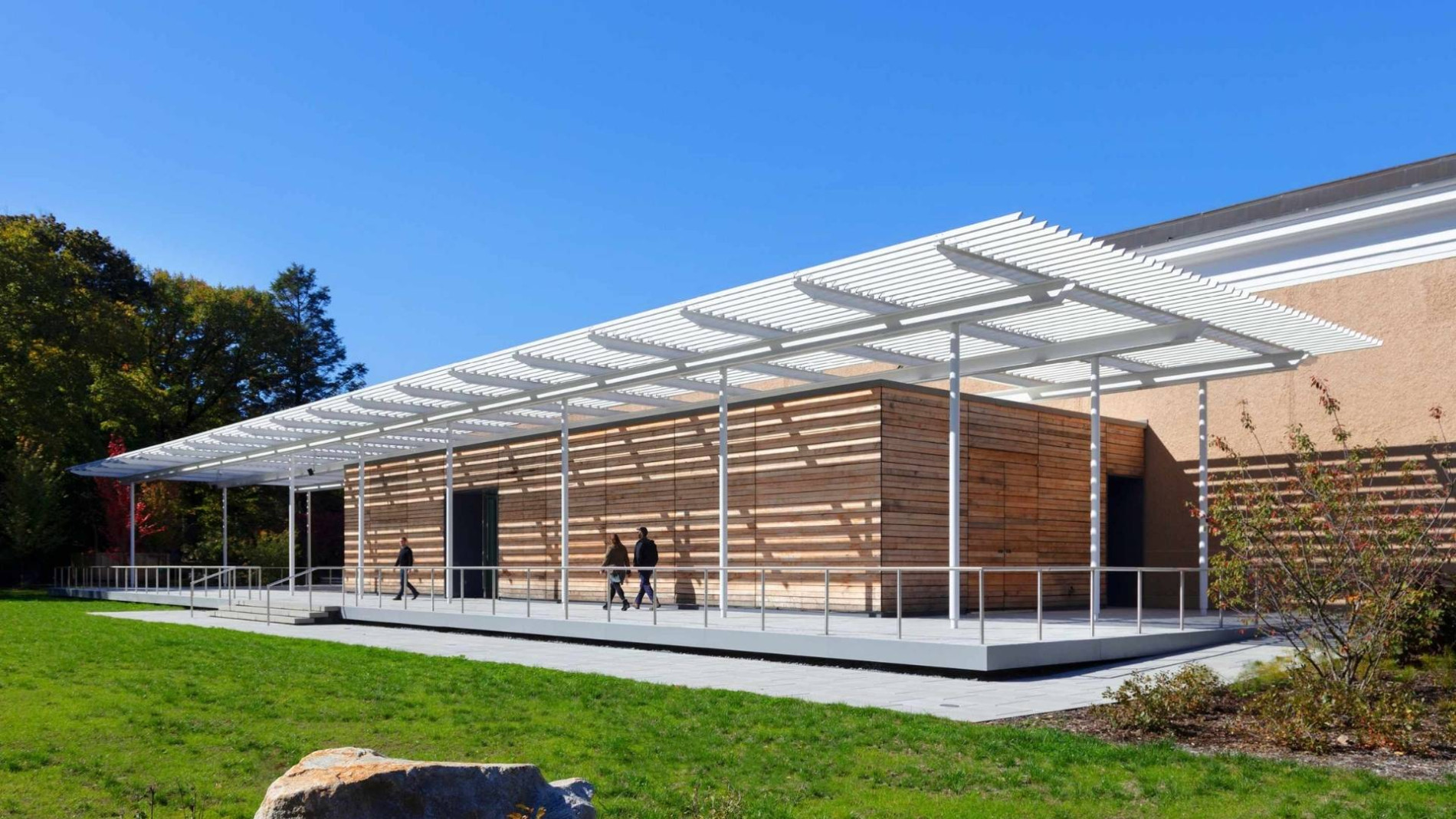 xterior view of the David Rockefeller Creative Arts Center, featuring a timber-clad building with a wide white canopy structure and people walking along the front pathway.