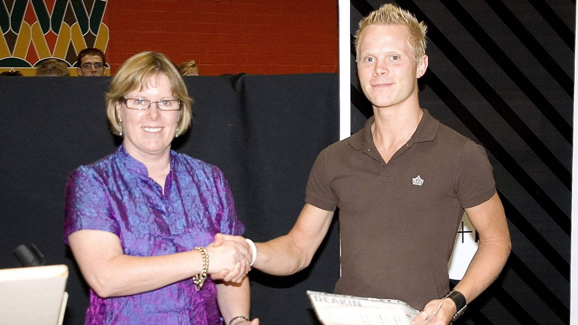 Martin shaking hands with a woman during an award or graduation ceremony, holding a certificate, with people watching in the background.