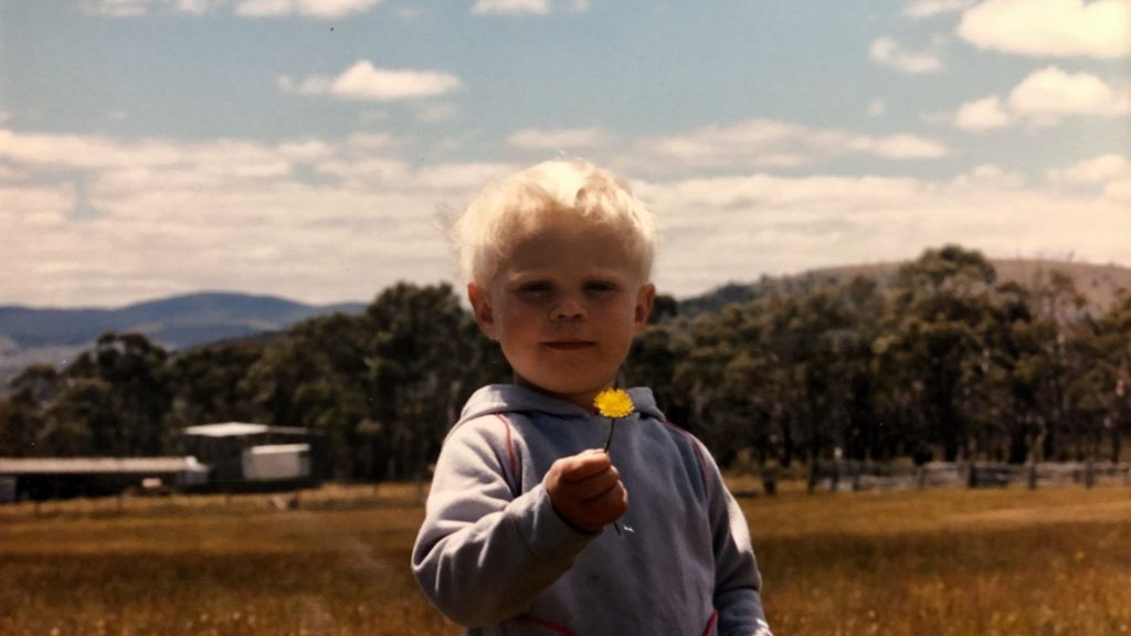 Young child with blonde hair holding a yellow flower in a field, with trees and hills in the background in rural Tasmania.