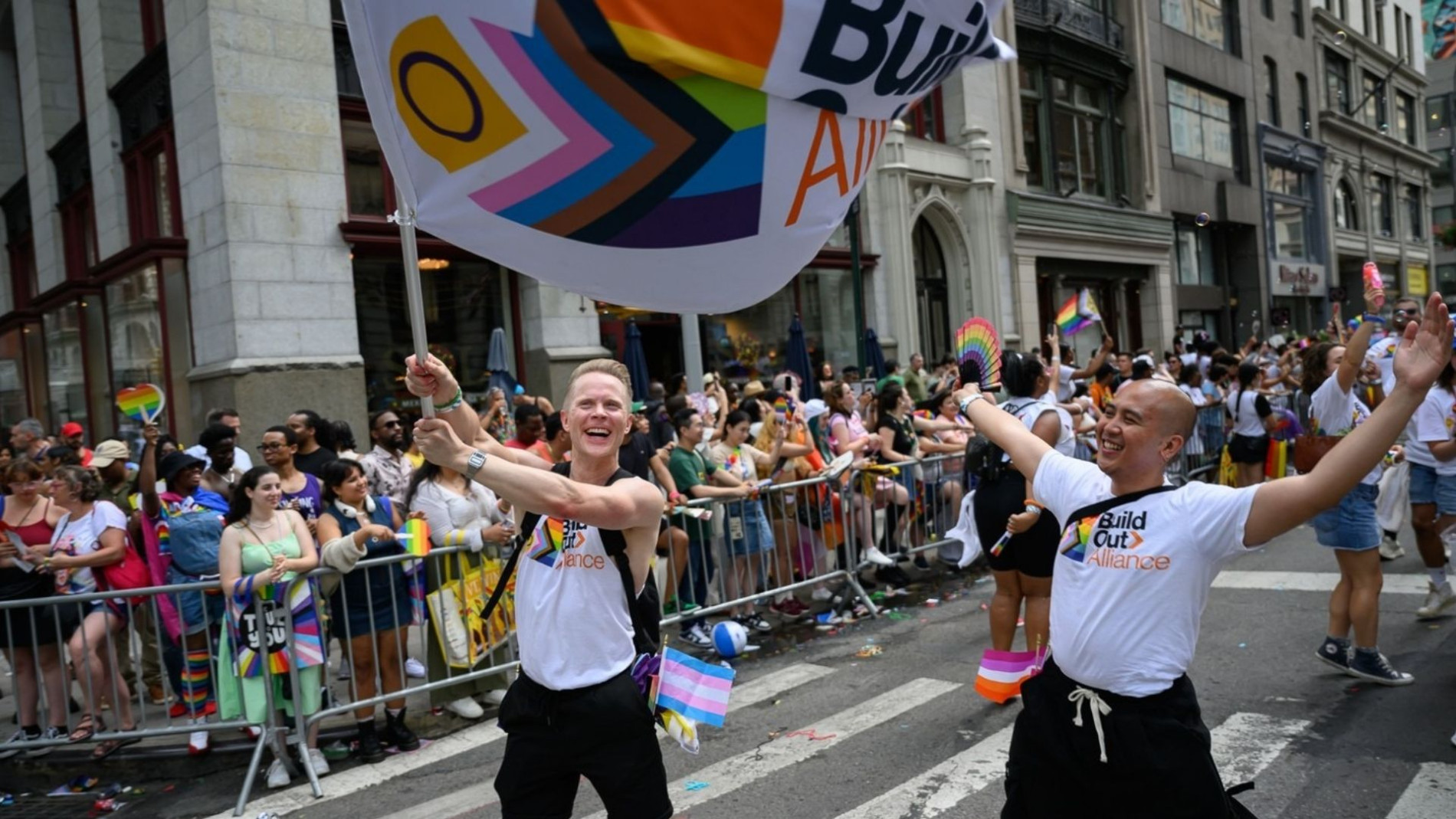Martin leading a Pride parade, smiling and celebrating while waving a large Build Out Alliance flag, with a cheering crowd behind barricades on a city street.