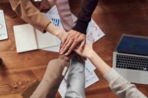 An image of work colleagues hands on top of each other over a desk. There are papers and a laptop on the desk to suggest a work environment. The hands suggest unity and teamwork.