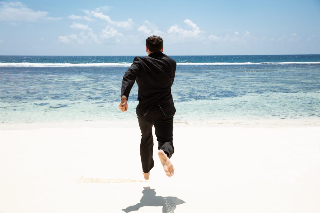 Rear View Of A Businessman Running Barefoot Toward The Idyllic Sea At Beach