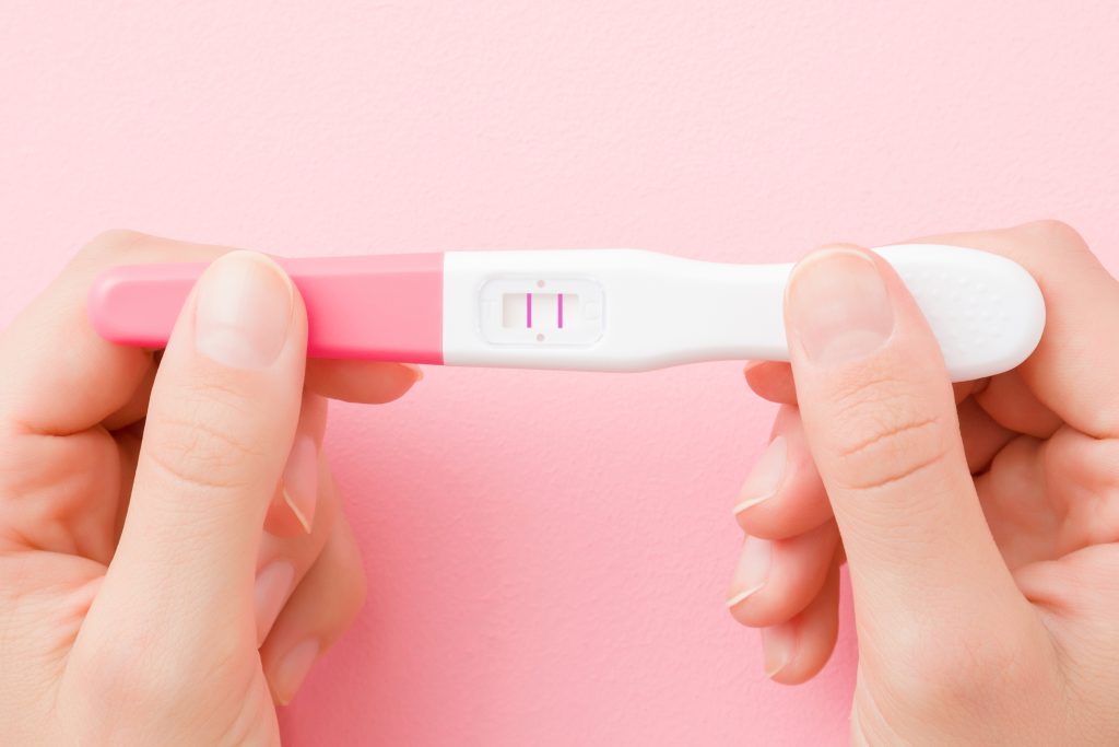 Young woman hands holding pregnancy test with two stripes on pastel pink background. Positive result. Closeup. Point of view shot. Top down view.