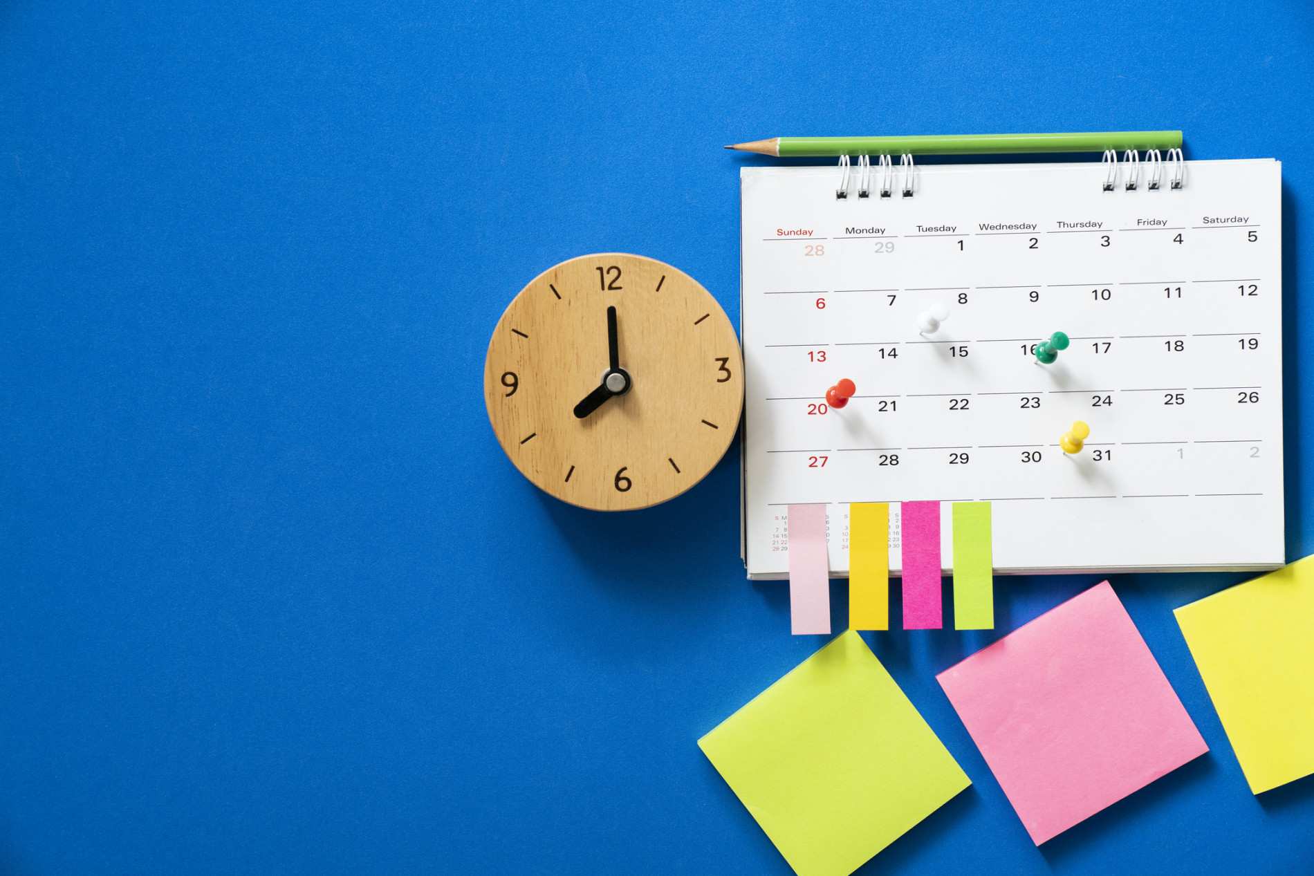 A blue background. An analog clock is placed next to a calendar, a pencil and lots of colourful post-it notes.
