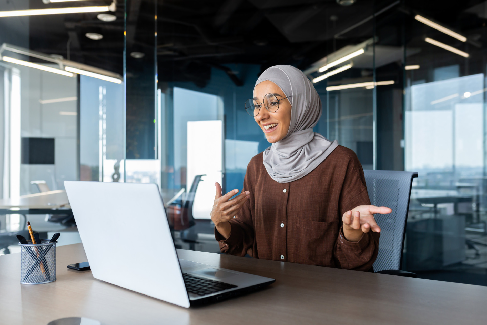 A woman wearing glasses and a hijab is sitting in front of a laptop. She is in an office environment. She is mid conversation, which suggests she is on a video call. 