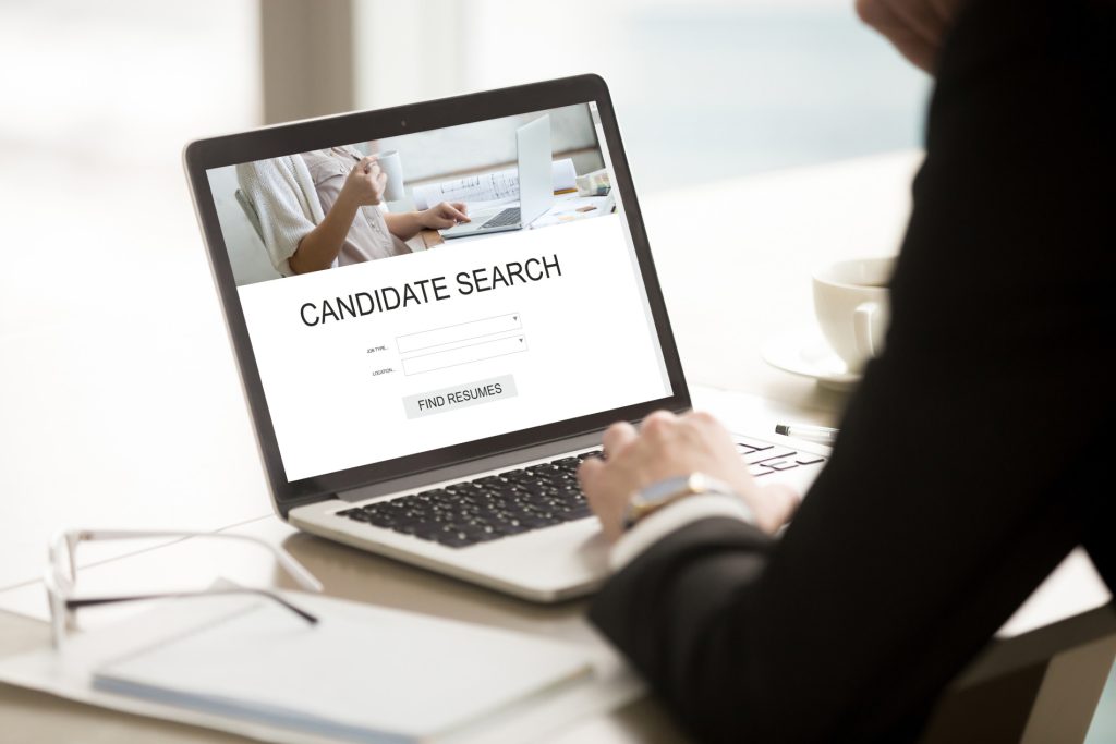 A laptop with the words 'candidate search' written on the screen. There is a person sat in front of the laptop with their hand on the keyboard. You can not see the person's face but it looks as if they are looking at the laptop screen, searching for candidates.