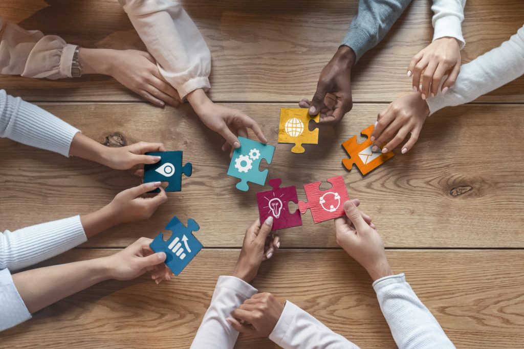 A team of diverse people sat around a table. You can only see there hands. They are all holding jigsaw pieces with various symbols on into the middle of the table. The image symbolises a team working together and sharing ideas.