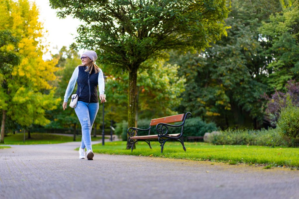A photo of a woman walking in a park. She is walking on a path, and there is a bench, grass and trees nearby. She looks happy and content.