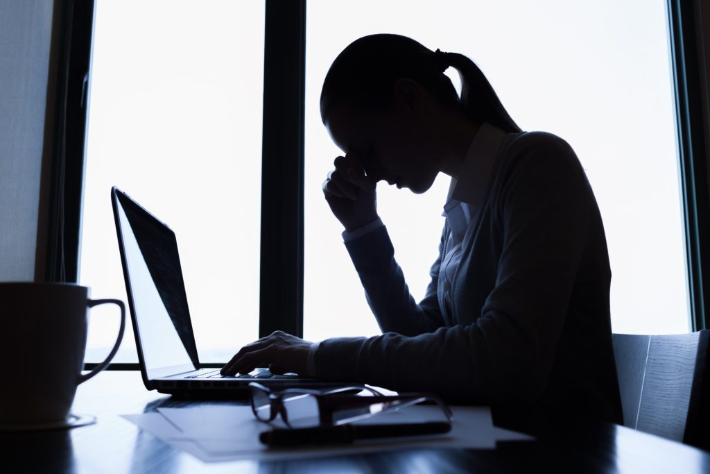 A woman sat at a desk looking at a laptop. The woman has her head in her hands, as if stressed. The photo is silhouetted to suggest the woman is sad or stressed.