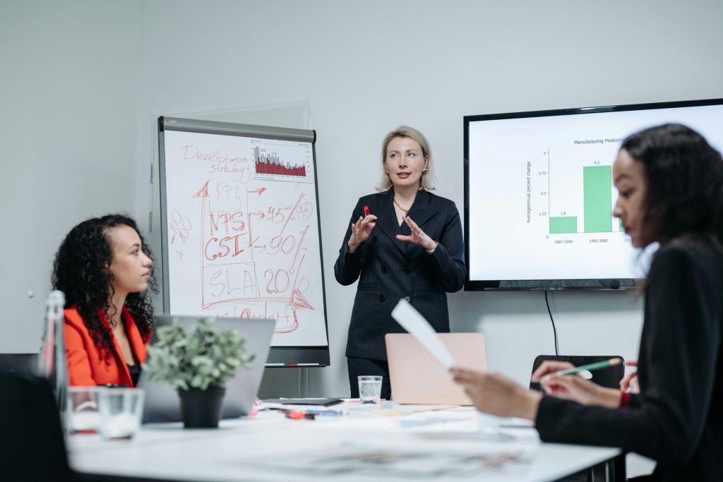 A woman presents business growth metrics to two colleagues in a professional meeting room, with a whiteboard and data screen behind her.