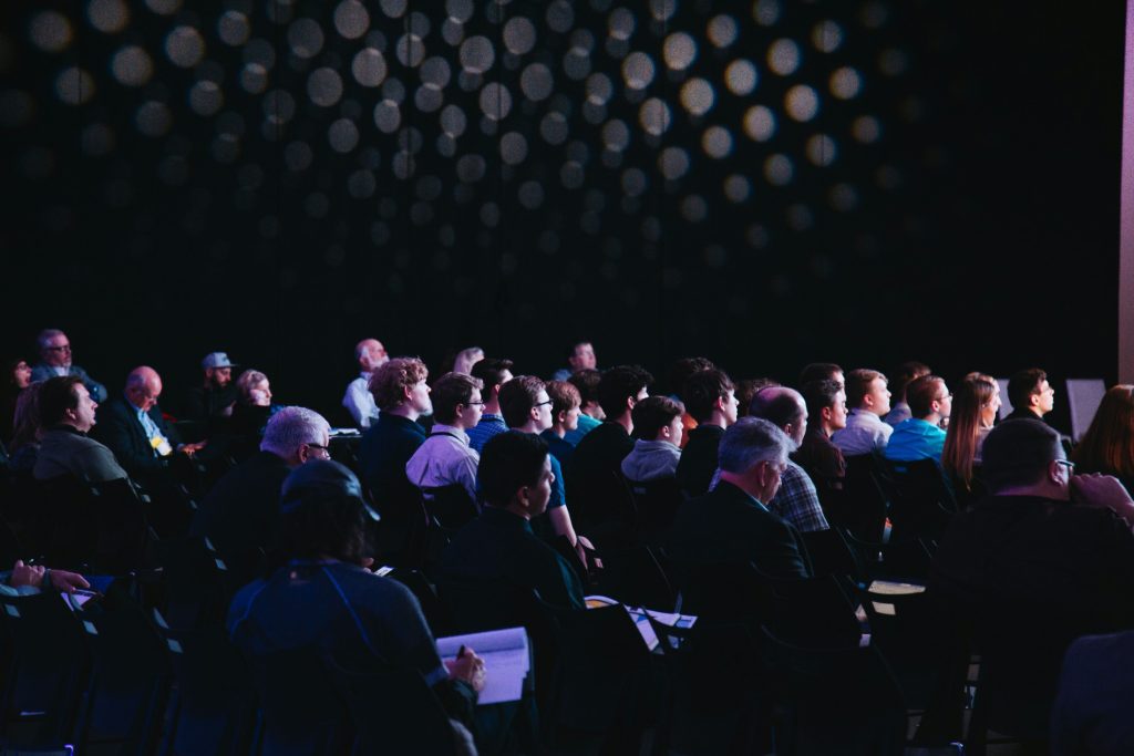 A seated audience at a B2B conference, engaged and taking notes, lit by warm stage lighting against a dark auditorium backdrop.