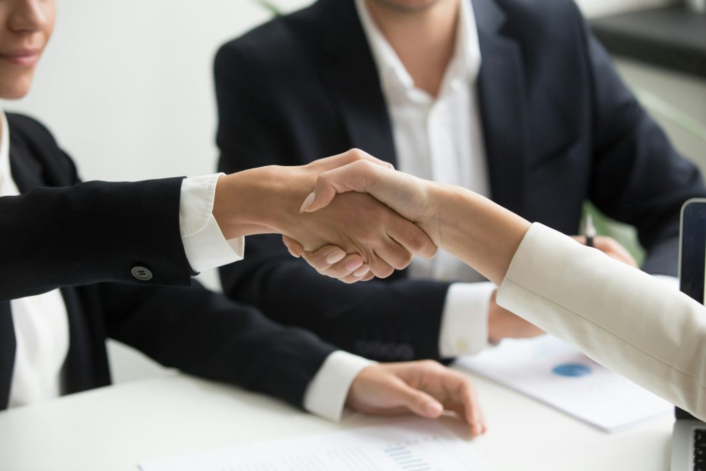 Two professionals shaking hands across a desk during a B2B events recruitment interview, with a third person seated in the background.