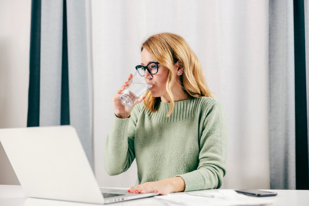 A woman drinking water while working remotely on her laptop from home office.